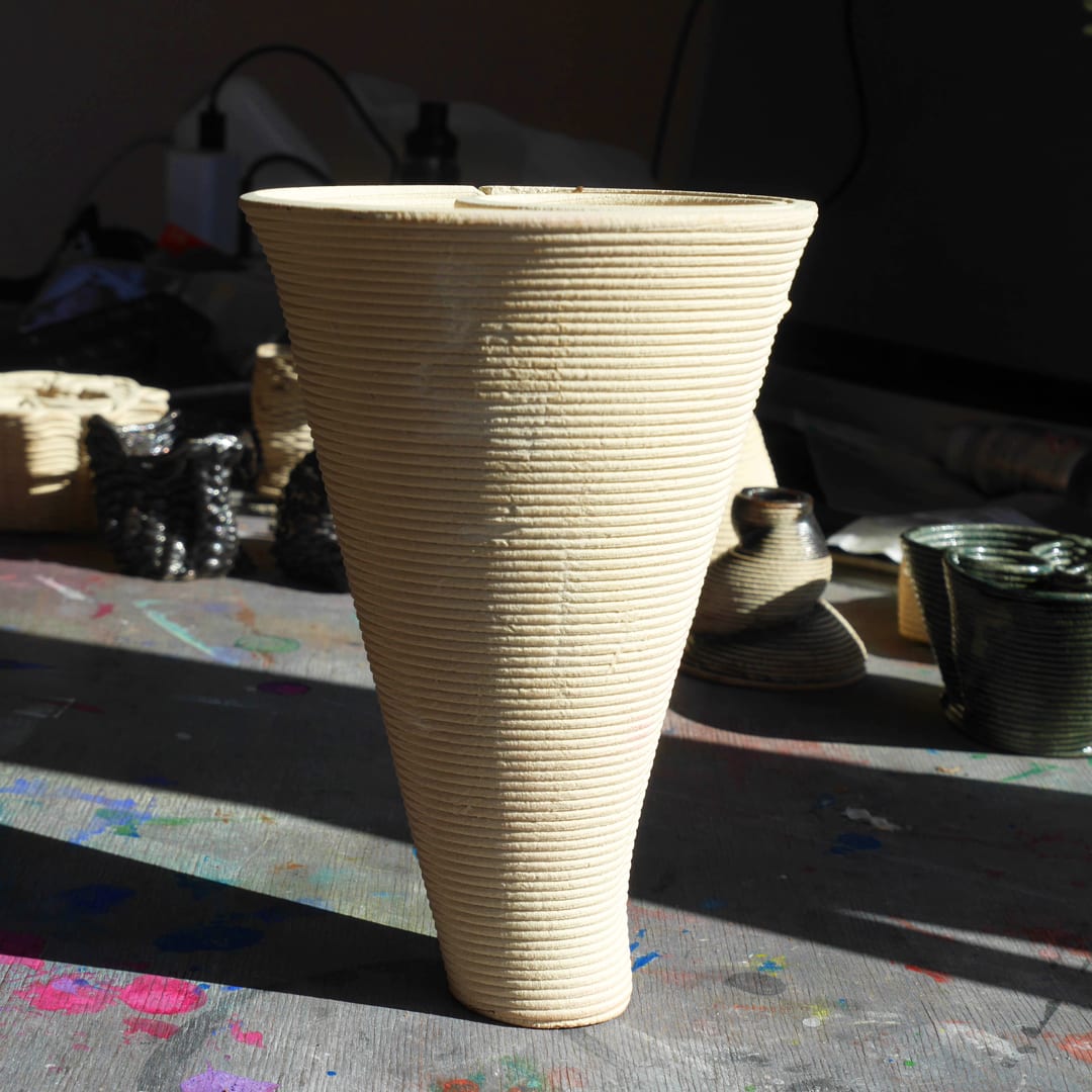 Tall conical vase in fine even coils, other pieces of the series behind it in shadow.